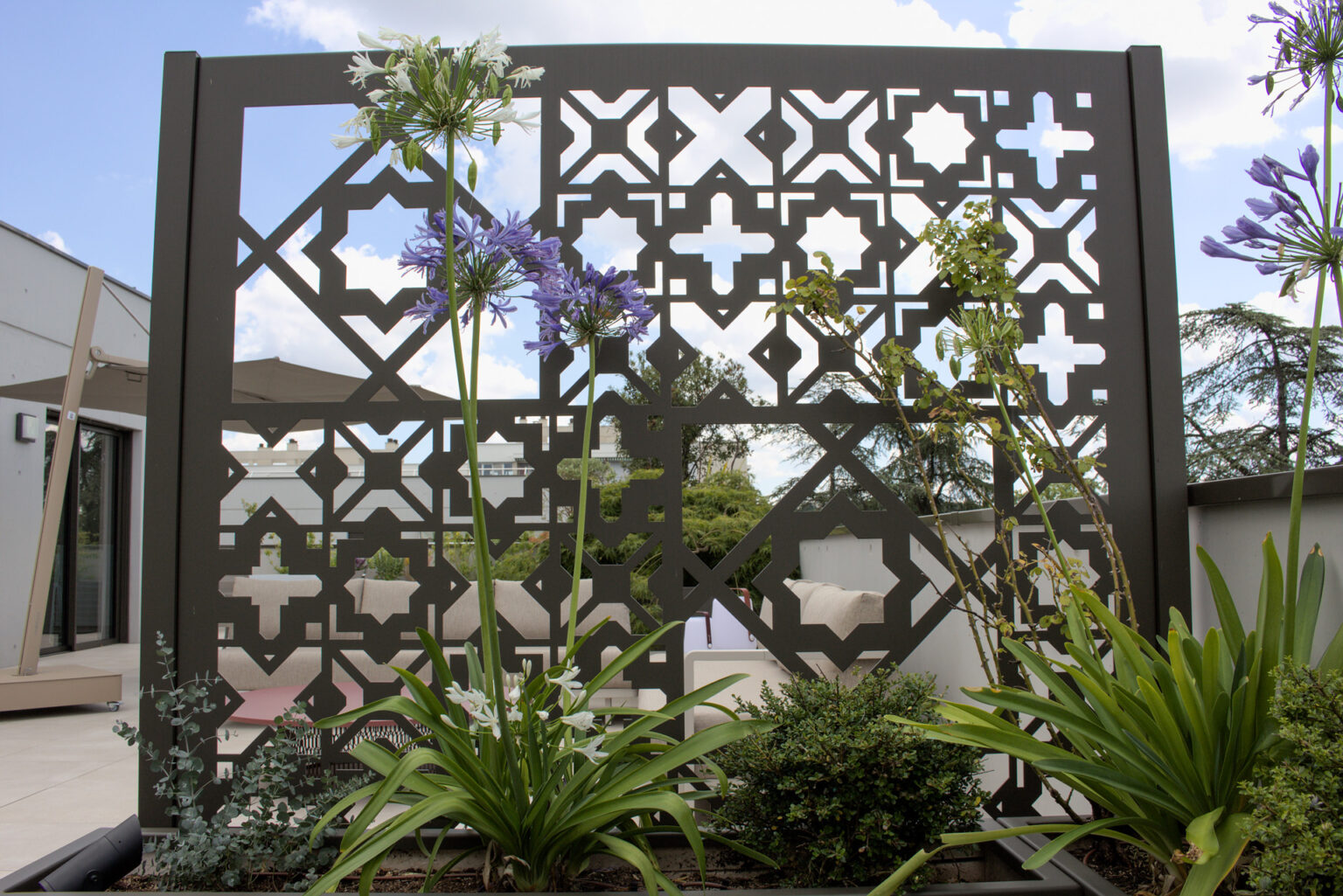 Claustra aux motifs géométriques en contre-jour, agapanthes bleues et blanches devant le salon – projet ACER