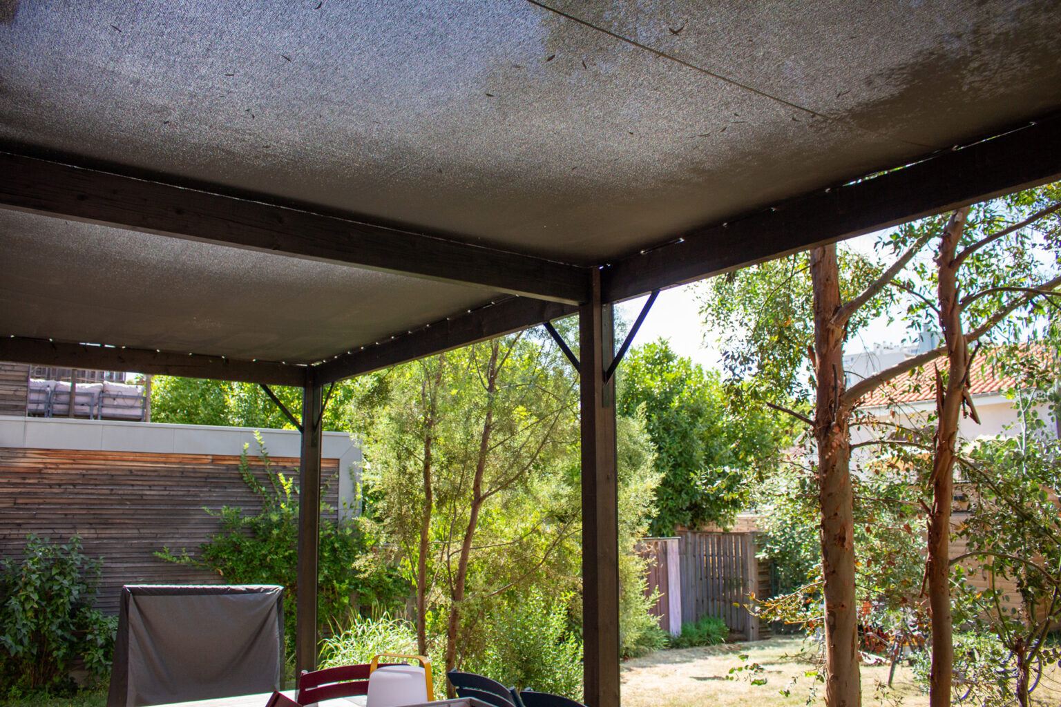 Sous la pergola bois en toile micro-perforée, vue sur le jardin, le bardage de la maison et la table – projet BETULA