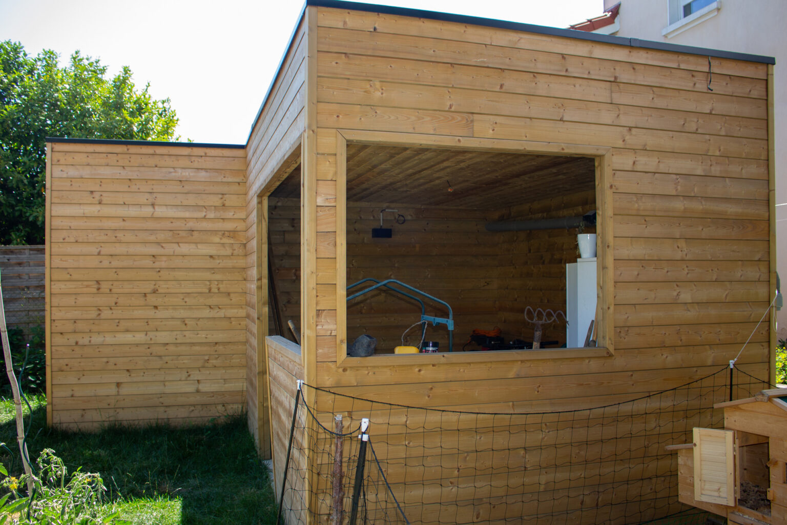 Abri de jardin bois à toit plat ouvert sur l'atelier de rangement, enclos à poules au premier plan – projet BETULA