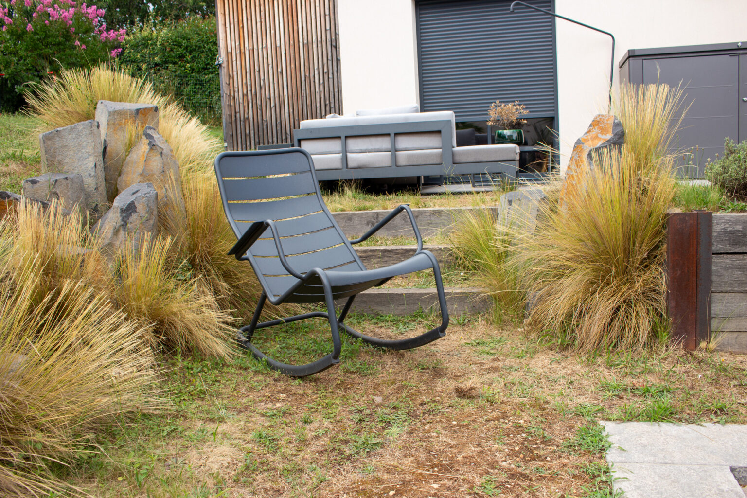 Fauteuil à bascule gris entre graminées et blocs de basalte, salon et façade bardée de bois en fond – projet BAOBAB