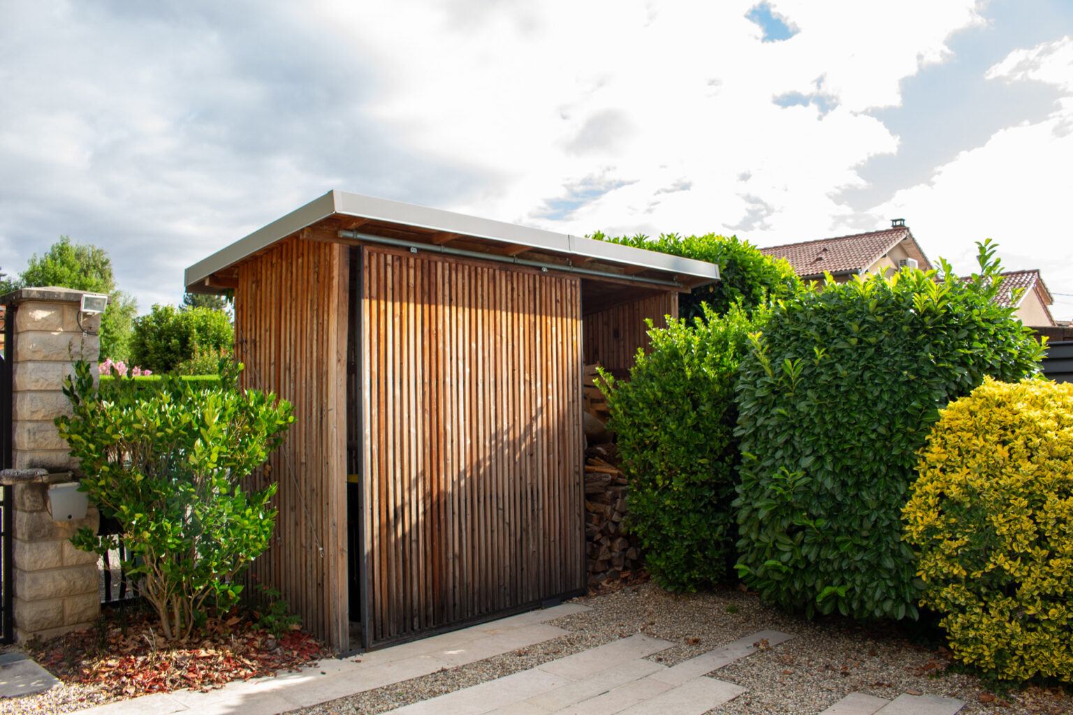 Abri de jardin en ossature bois habillé de tasseaux, porte coulissante ouverte sur le rangement à bûches – projet CAMELLIA