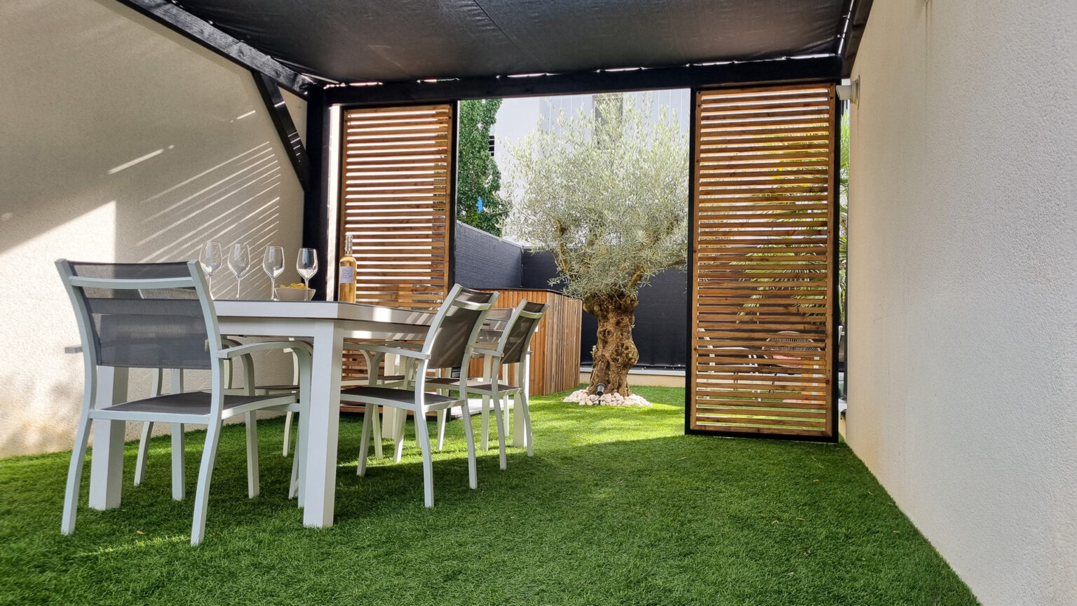 Table à manger sous la pergola entre deux panneaux coulissants en tasseaux mélèze, olivier au fond – projet GAURA