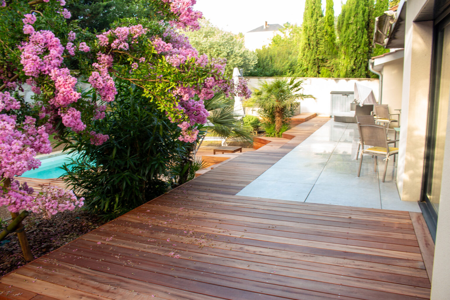 Lagerstroemia rose au-dessus du cheminement en bois exotique reliant la terrasse grès cérame à la piscine – projet LAVANDULA