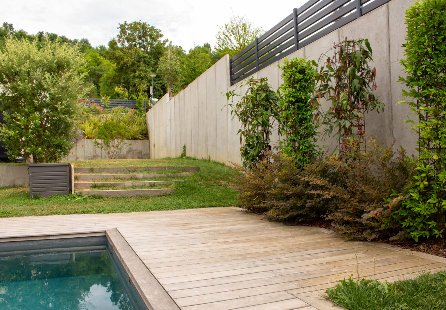 Piscine avec plage en bois, pelouse, marches en bois et mur de soutènement en béton banché végétalisé – projet LOROPETALUM