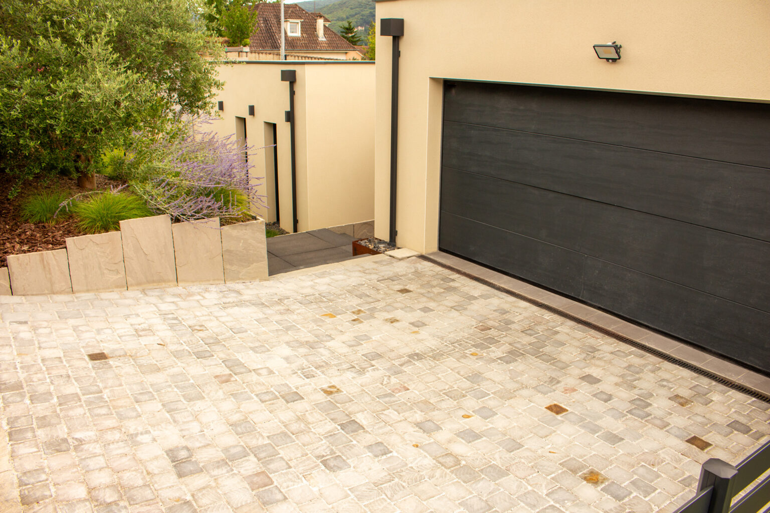 Cour en pavés de grès gris devant une porte de garage noire, massif retenu par des plaques de grès – projet LOROPETALUM