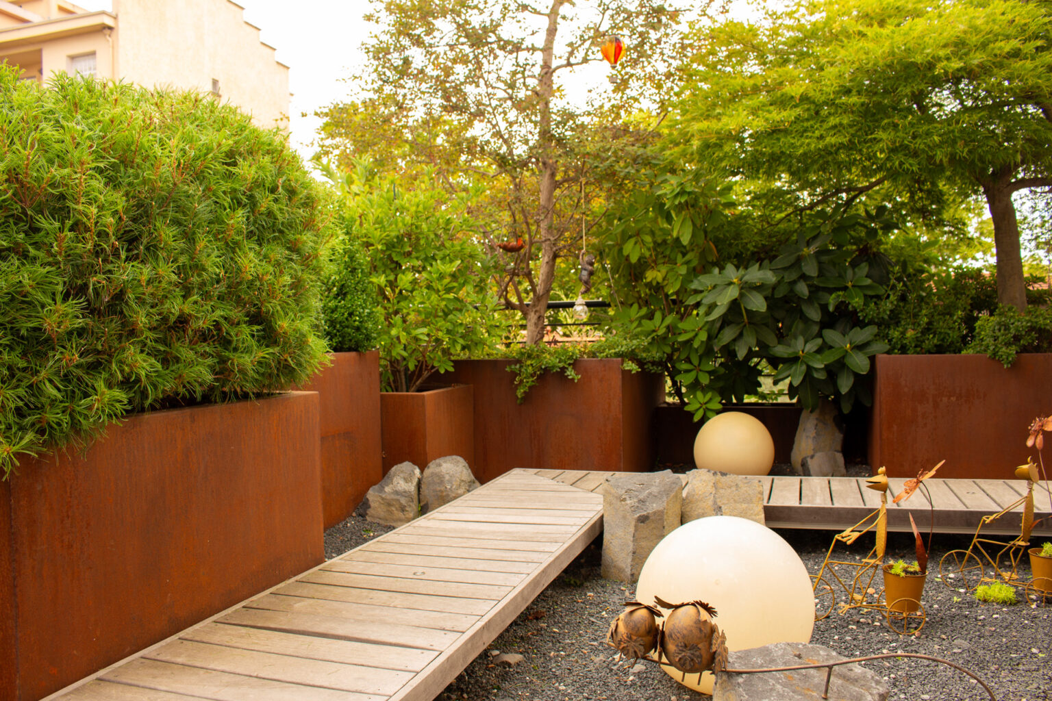 Pontons en ipé, rochers et boules lumineuses sur gravier entre des bacs en corten plantés d'arbustes – projet PAEONIA