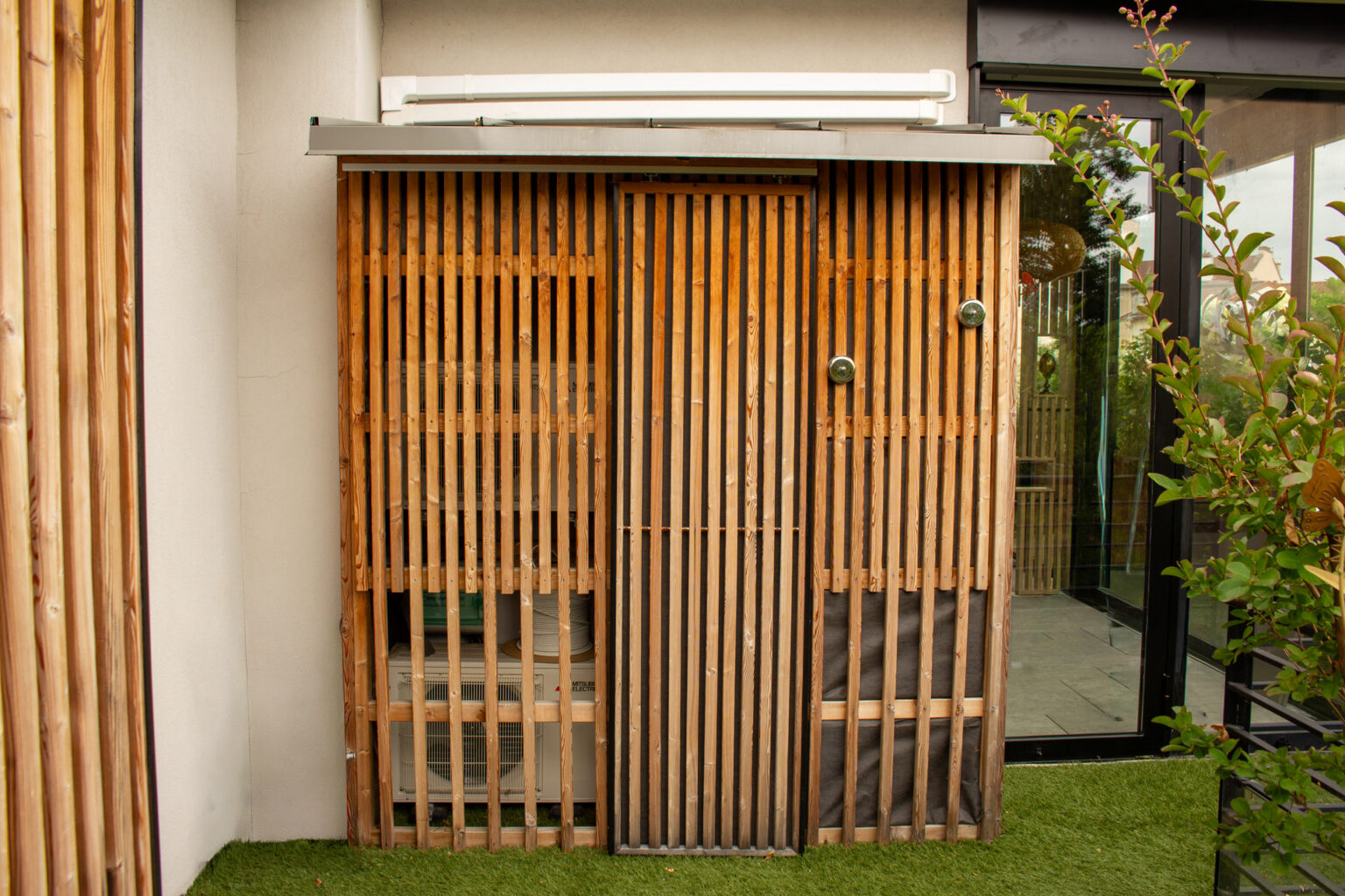 Rangement en bois à claire-voie abritant la pompe à chaleur, porte coulissante et gazon synthétique – projet PAEONIA