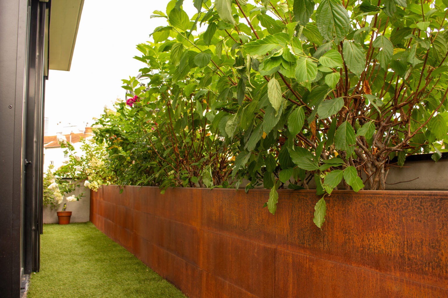 Jardinière en acier corten fabriquée sur place, hortensias et gazon synthétique le long de la façade – projet PAEONIA