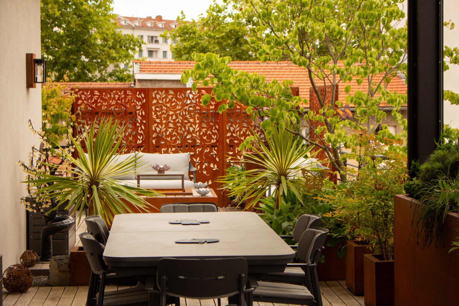 Grande table à manger sur ponton en ipé devant des claustras en corten ajouré, cordylines et salon cosy – projet PAEONIA