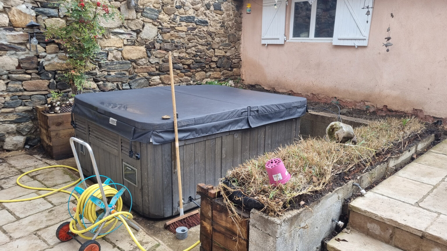 Avant travaux : spa bâché sur dalles devant un mur de pierre, jardinière en parpaings délabrée à côté – projet SYRINGA