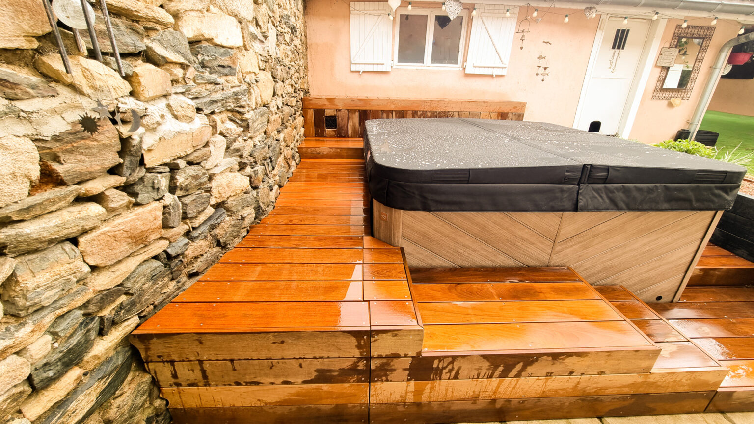 Plage en ipé sur deux niveaux longeant un mur de pierre, spa couvert et banquette bois au fond – projet SYRINGA