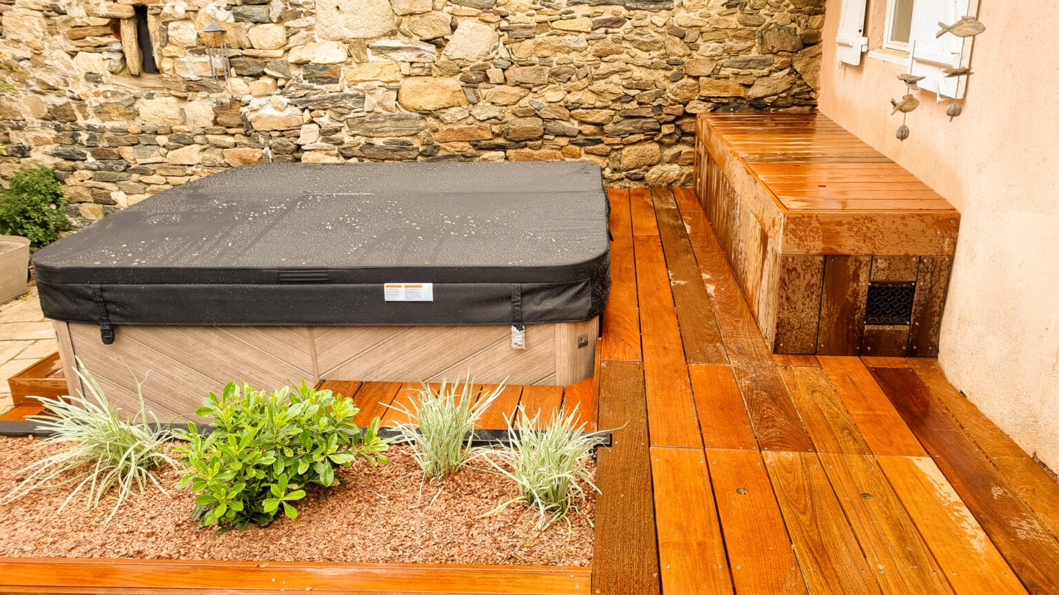 Banquette-coffre en bois exotique contre la façade, plage ipé et spa habillé au pied d'un mur de pierre – projet SYRINGA