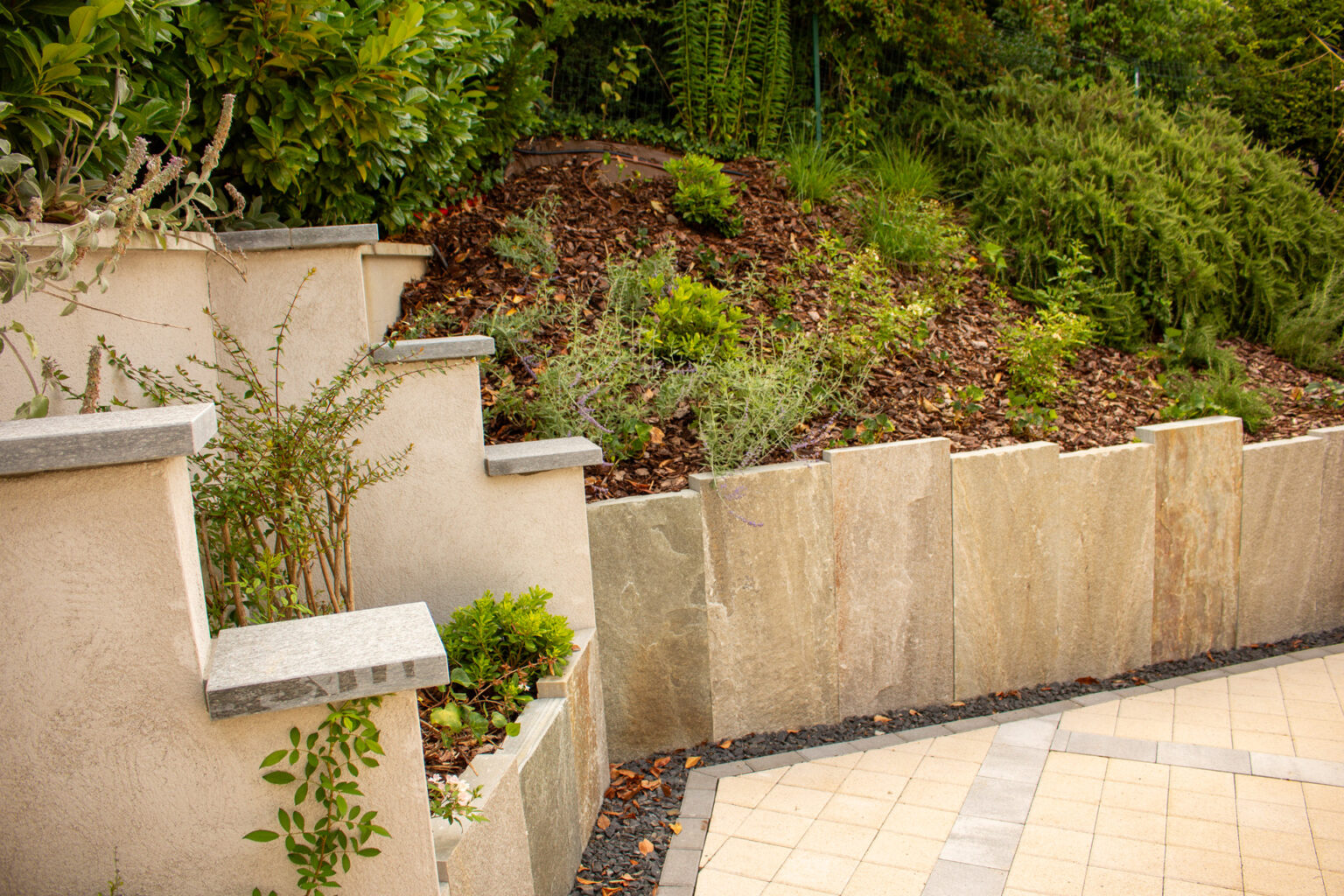 Talus paillé planté de vivaces au-dessus d'un mur en plaques minérales, concassé noir et pavage beige – projet ULMUS