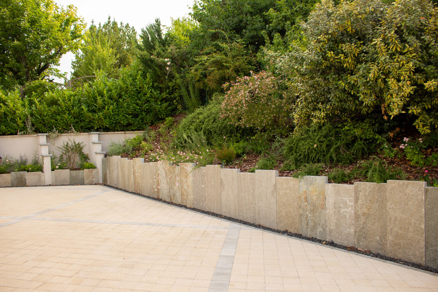 Cour pavée beige en pavés drainants longée par un mur de soutènement en plaques minérales et un talus planté – projet ULMUS