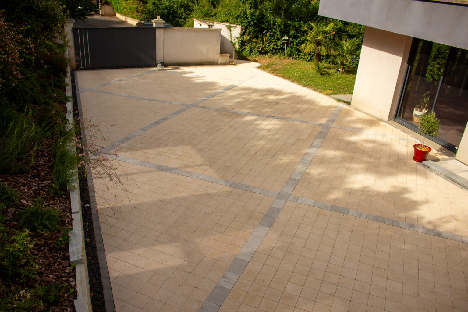 Cour d'accès en pavés drainants beiges traversée de lignes de pavés gris, du portail coulissant vers la maison – projet ULMUS
