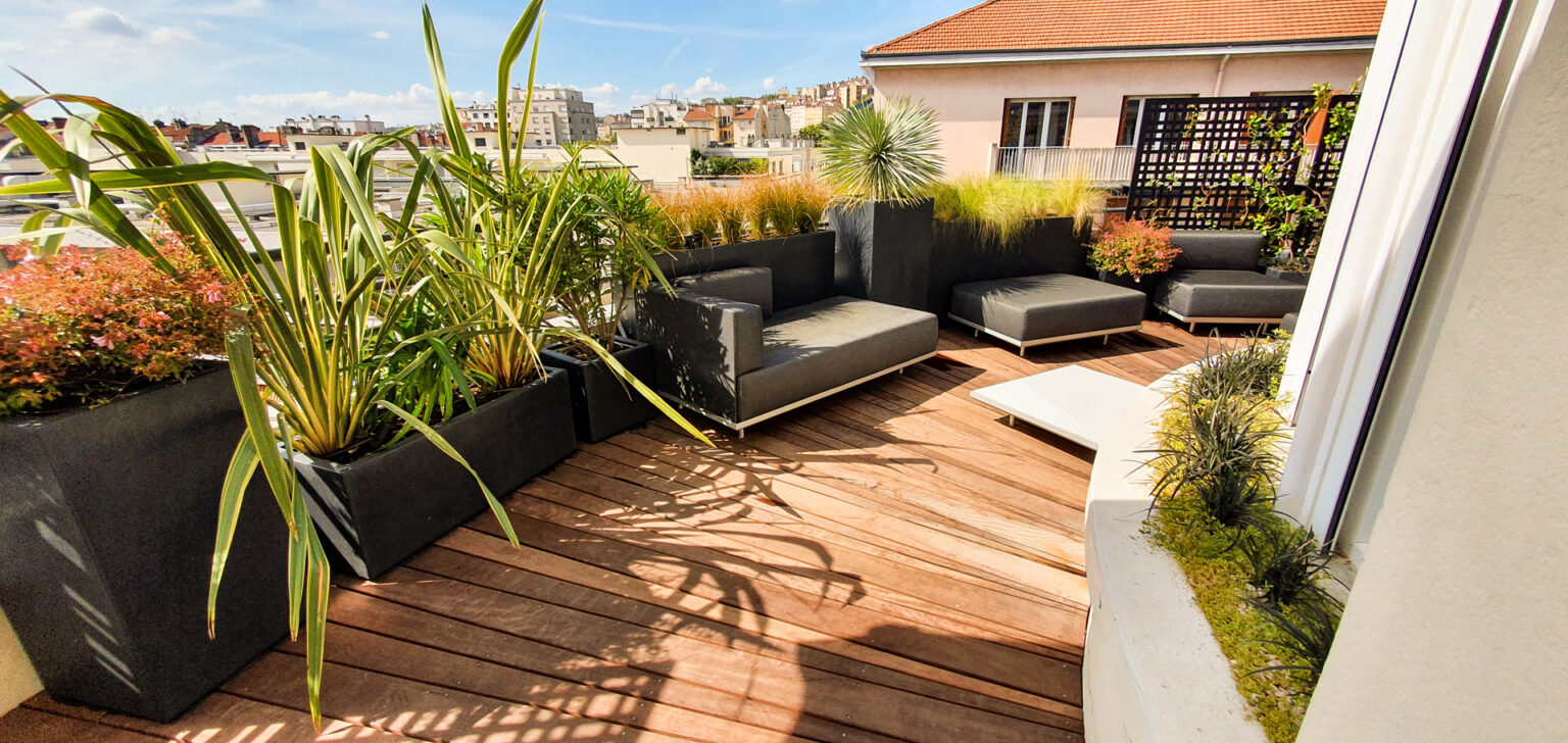 Salon lounge gris sur platelage bois rayonnant d'une terrasse arrondie, phormiums en bacs au premier plan – projet SEDUM