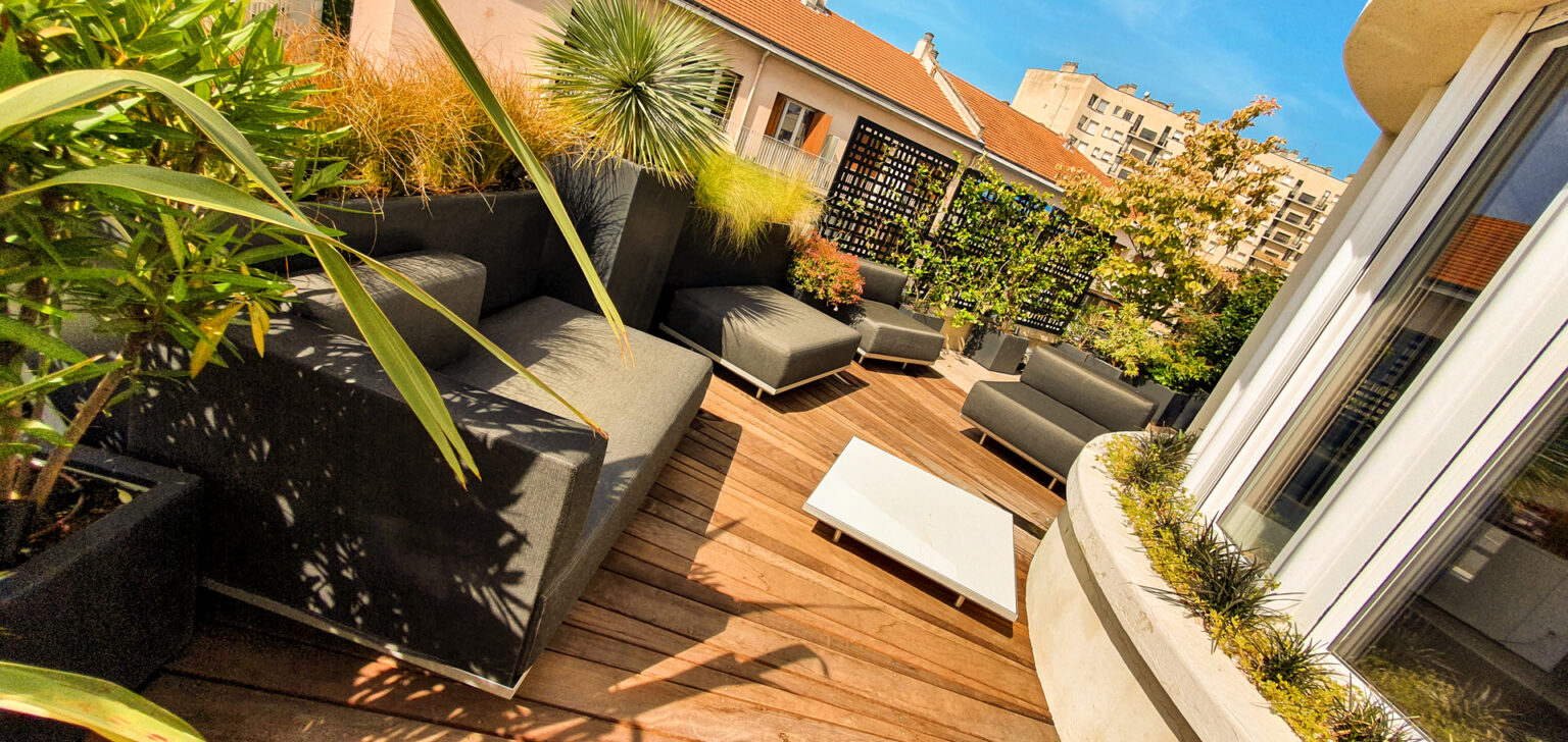 Terrasse arrondie en lames bois posées en éventail, canapés d'angle gris et bacs anthracite de graminées – projet SEDUM