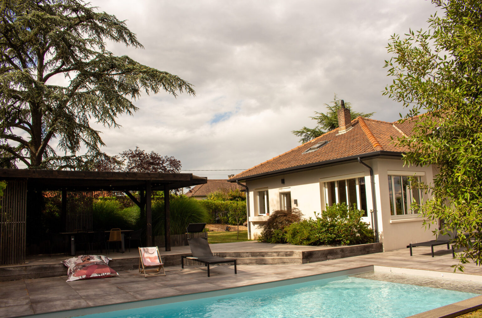 Pergola bois, terrasse dallée et piscine devant la maison sous un ciel nuageux, transat et coussins de sol – projet SALVIA