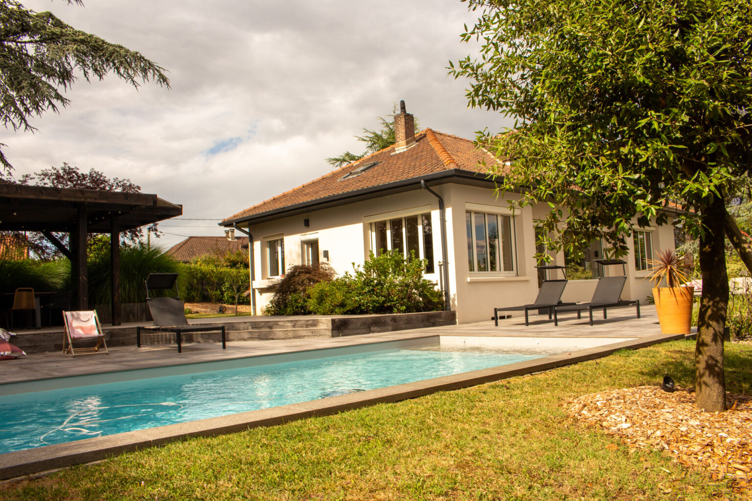 Bassin de piscine étiré entre pelouse et terrasse dallée, pergola bois et bains de soleil devant la maison – projet SALVIA