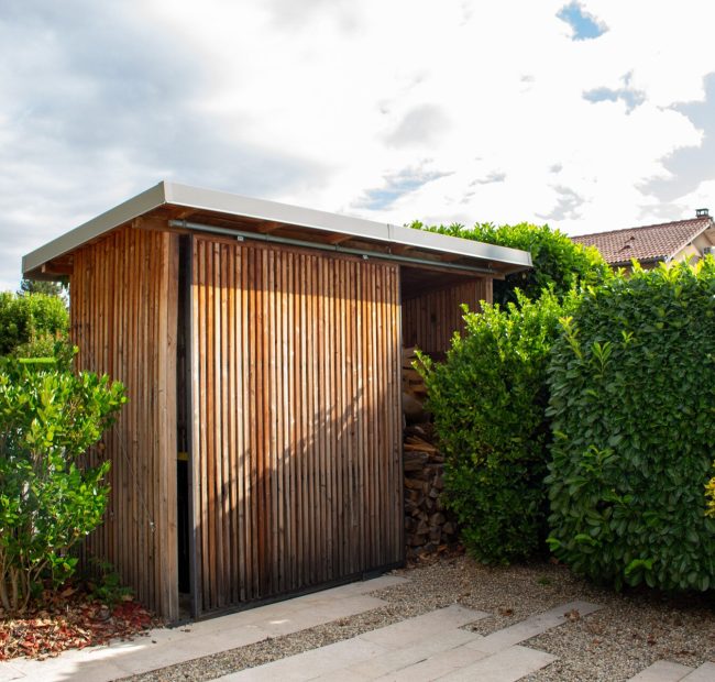 Abri de jardin en ossature bois habillé de tasseaux, porte coulissante ouverte sur le rangement à bûches – projet CAMELLIA